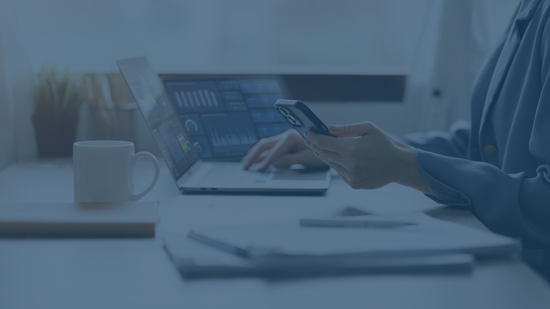 Eine Person sitzt an einem Schreibtisch und arbeitet an einem Laptop, der mehrere Dashboards mit Diagrammen und Statistiken anzeigt. In der Hand hält sie ein Smartphone. Auf dem Schreibtisch stehen eine weiße Tasse sowie ein Notizbuch, und im Hintergrund ist eine helle, moderne Büroumgebung zu erkennen. Das Bild vermittelt einen professionellen Eindruck, passend zum Thema der effizienten plattformübergreifenden Geräteverwaltung mit Microsoft Intune.