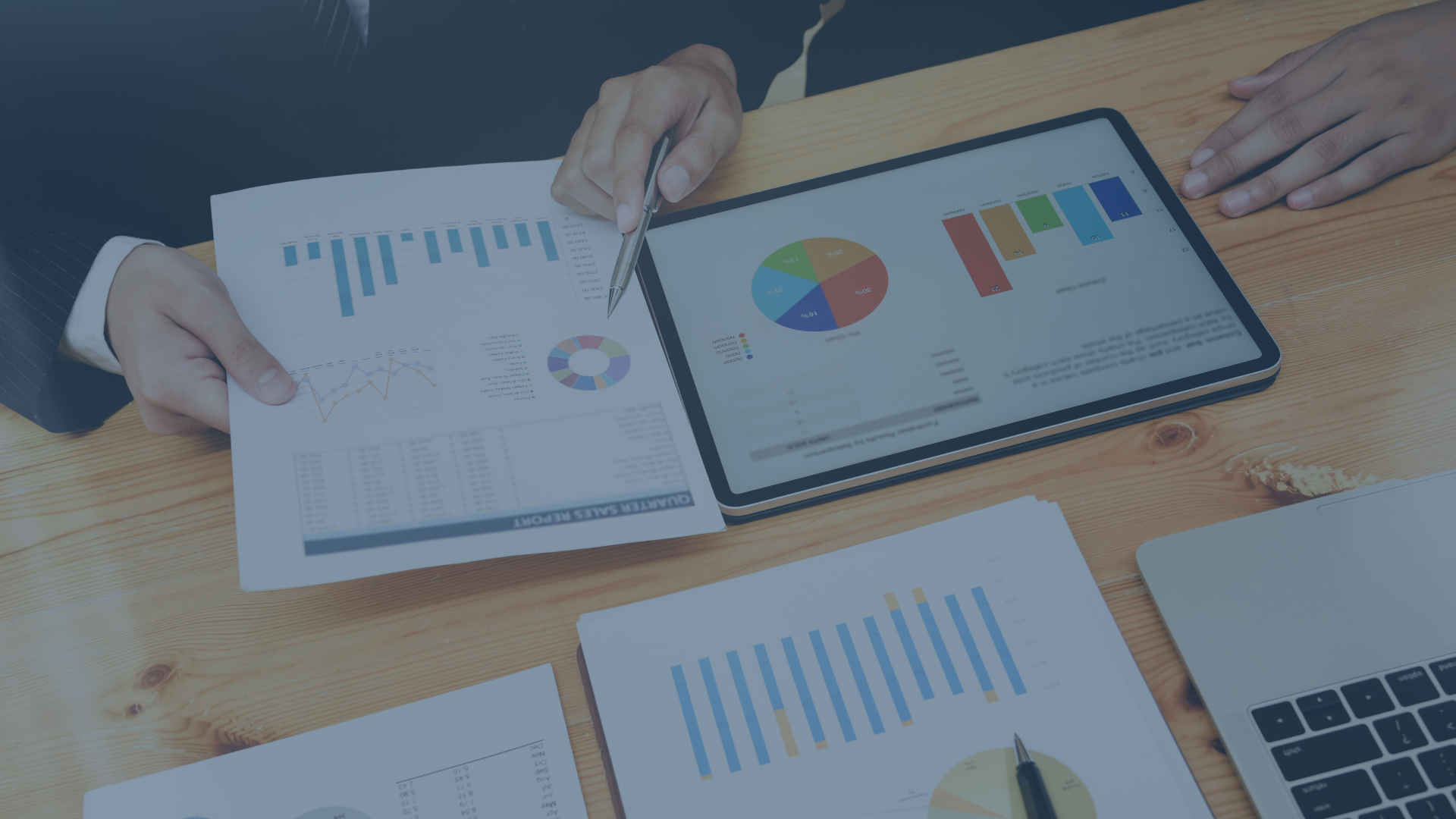 Das Bild zeigt zwei Personen an einem Holztisch, die gemeinsam Geschäftsdaten analysieren. Auf dem Tisch liegen mehrere Diagramme und Berichte mit Balken-, Linien- und Kreisdiagrammen. Eine Person hält ein Blatt mit einem Verkaufsbericht, während die andere auf ein Tablet zeigt, das farbige Grafiken darstellt. Ein Laptop ist ebenfalls zu sehen. Die Szene vermittelt eine professionelle Besprechung oder Datenanalyse im geschäftlichen Kontext.