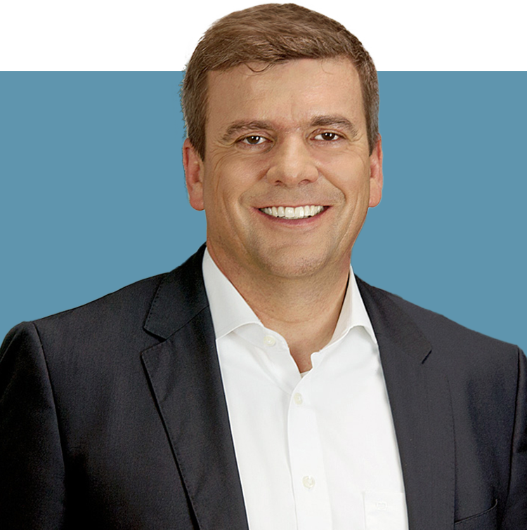 Porträt von Rainer Francisi, Vorstandsmitglied der audius SE, lächelnd in dunklem Sakko und weißem Hemd vor blauem Hintergrund.