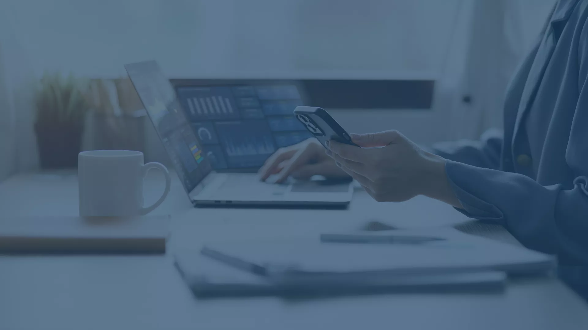 Eine Person sitzt an einem Schreibtisch und arbeitet an einem Laptop, der mehrere Dashboards mit Diagrammen und Statistiken anzeigt. In der Hand hält sie ein Smartphone. Auf dem Schreibtisch stehen eine weiße Tasse sowie ein Notizbuch, und im Hintergrund ist eine helle, moderne Büroumgebung zu erkennen. Das Bild vermittelt einen professionellen Eindruck, passend zum Thema der effizienten plattformübergreifenden Geräteverwaltung mit Microsoft Intune.