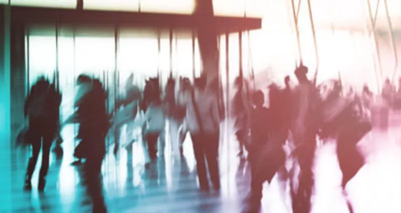 Verschwommenes Foto einer Menschenmenge, die durch einen hellen, modernen Raum mit Glaswänden und Sonnenlicht geht. Die Menschen sind in Bewegung und ihre Umrisse sind unscharf, was ein Gefühl von Hektik und Aktivität vermittelt.