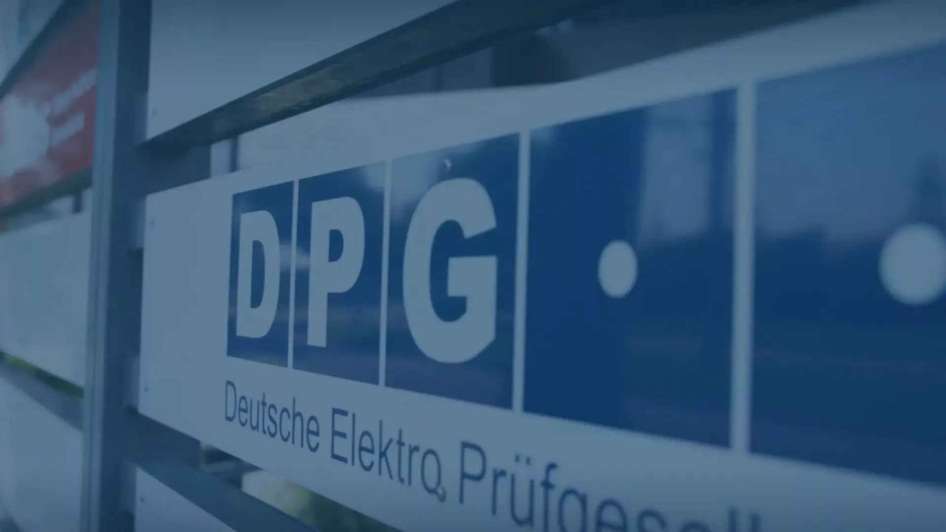 Nahaufnahme des blauen Firmenlogos der DPG Deutsche Elektro Prüfgesellschaft an einem Zaun oder einer Außenfassade eines Firmengebäudes. Das Schild zeigt die Initialen D P G mit einem Punkt und den darunter stehenden Schriftzug "Deutsche Elektro Prüfgesell".