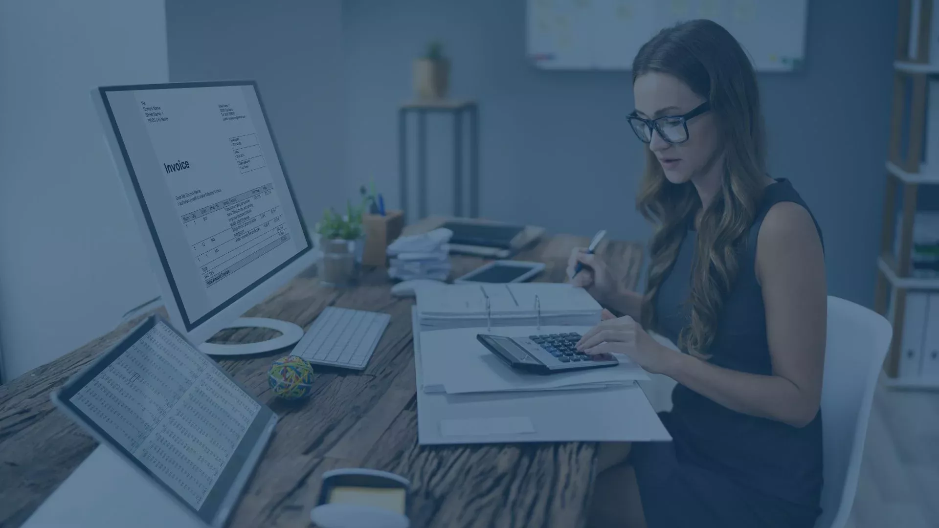 Buchhalterin arbeitet im Büro mit Taschenrechner, Unterlagen und zwei Monitoren, auf denen Rechnungen und Tabellen angezeigt werden.