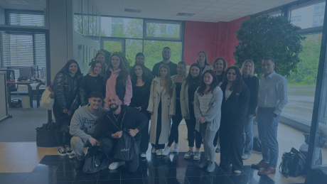 Eine größere Gruppe von Schülerinnen, Schülern und Mitarbeitenden steht gemeinsam in einem modernen Eingangsbereich eines Unternehmens. Alle blicken lächelnd in die Kamera. Im Hintergrund sind große Fenster, Büroräume und Pflanzen zu sehen.