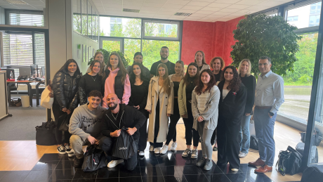 Eine größere Gruppe von Schülerinnen, Schülern und Mitarbeitenden steht gemeinsam in einem modernen Eingangsbereich eines Unternehmens. Alle blicken lächelnd in die Kamera. Im Hintergrund sind große Fenster, Büroräume und Pflanzen zu sehen.