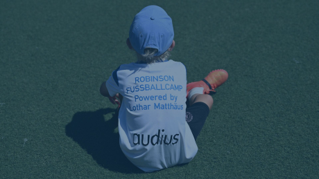 Ein Kind sitzt in Fußballkleidung auf einem Kunstrasenplatz, trägt ein Trikot mit der Aufschrift „Robinson Fußballcamp powered by Lothar Matthäus“ sowie dem Sponsorenlogo „audius“. Das Bild vermittelt die Idee der Nachwuchsförderung und des Teamgeists im Rahmen des Fußballcamps auf Kos, das jungen Sporttalenten sportliche Entwicklung und Begeisterung für Fußball ermöglicht.